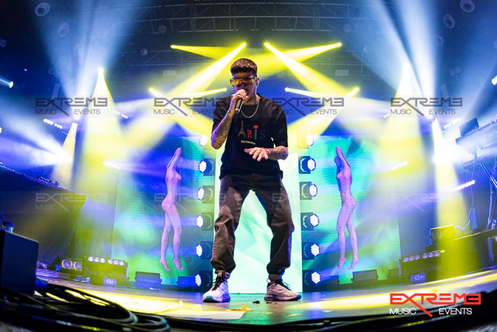 Maikel De la Calle performing live on stage wearing oversized white t-shirt with colorful lights in the background